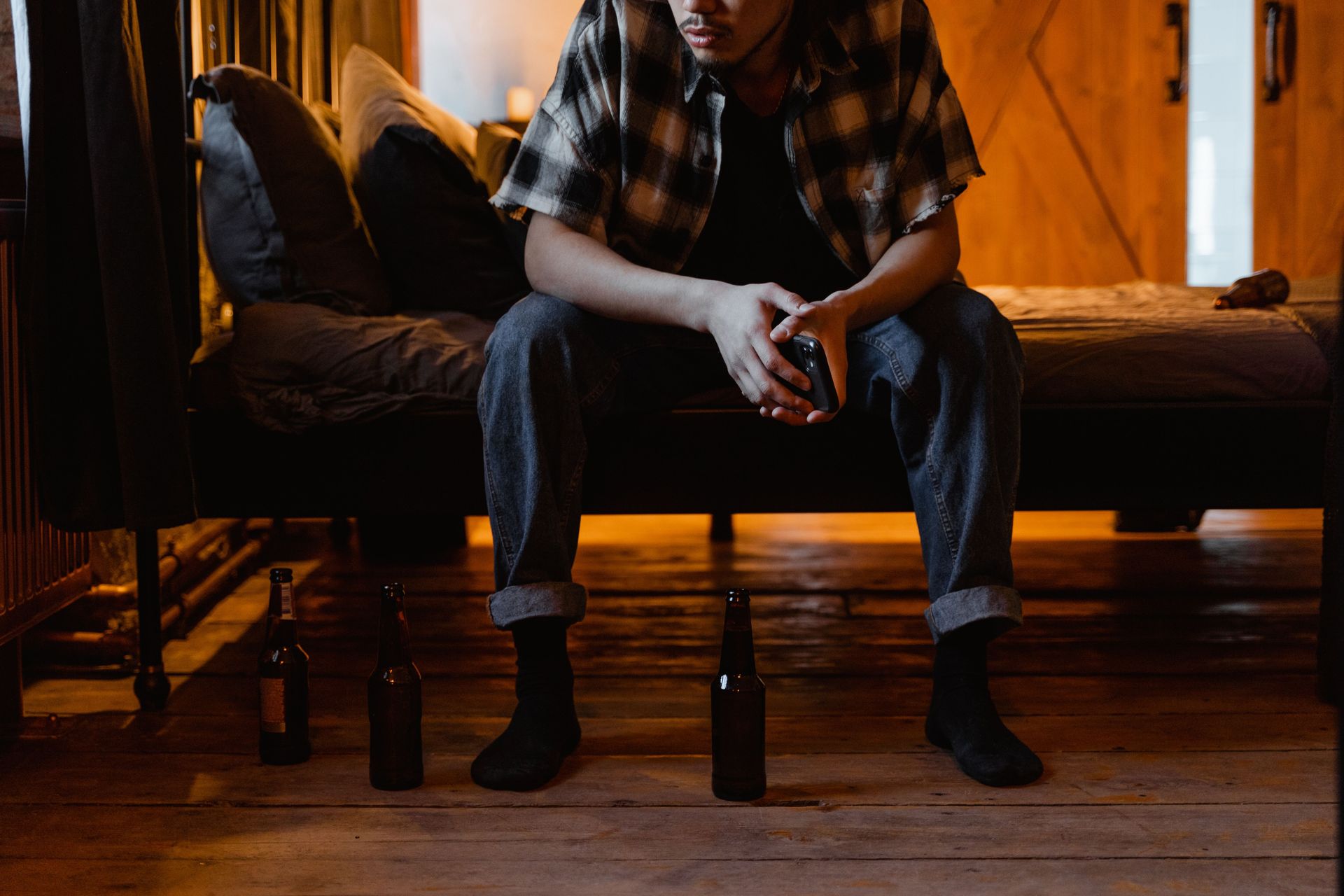 A man sits on a bed holding a cellphone, surrounded by beer bottles, creating a moody atmosphere.